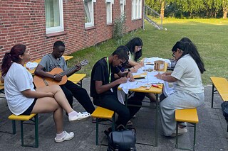 Jugendliche sitzen an Biertischen, einige bemalen T-Shirts, jemand spielt auf der Gitarre.