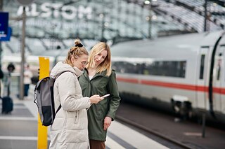 Zwei blonde Frauen stehen in einer Bahnhofshalle und blicken auf ein Smartphone. Im Hintergrund sieht man einen ICE.