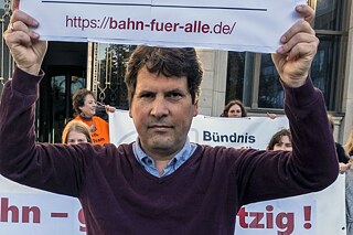 Ein Mann, Carl Waßmuth, hält ein Schild in die Luft, auf dem steht: „https://bahn-fuer-alle.de“. Im Hintergrund sieht man Demonstrierende mit Plakaten und Bannern.