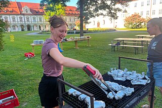 Eine junge Frau steht vor einem großen Grill, auf dem in Alufolie gewickeltes Gargut liegt.
