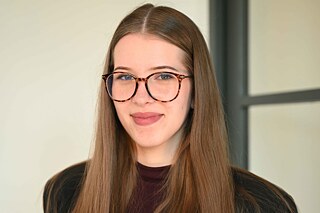 Eine junge Frau mit langem Haar lächelt. Sie trägt eine große Brille.