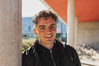 Ein junger Mann mit lockigem Haar leht an einer Säule und lächelt in die Kamera.