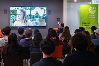 Schülerinnen und Schüler schauen auf einer Leinwand einen Film. Vor ihnen sitzt der Regisseur.