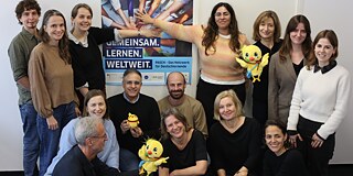 PASCH-Mitarbeitende stehen vor einem PASCH-Plakat. Auf dem PASCH-Paklat steht der Slogan: Gemeinsam.Lernen.Weltweit.