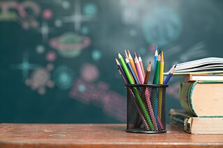 Stifte und Schulbücher liegen auf einem Tisch. Im Hintergrund eine bemalte, unscharfe Tafel.