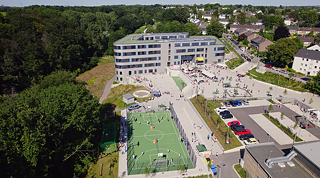 Schulgelände mit Gebäude, Sportplatz, Parkplätzen und umliegenden Wäldern und Straßen