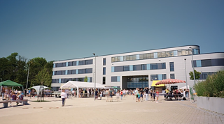 Modernes Schulgebäude, davor vier Stände und Menschen