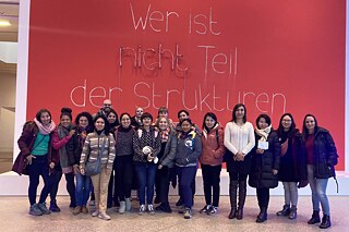 Eine Gruppe Erwachsener in der Bundeskunsthalle Bonn vor einer roten Wand auf der steht: Wer ist nicht Teil der Strukturen...