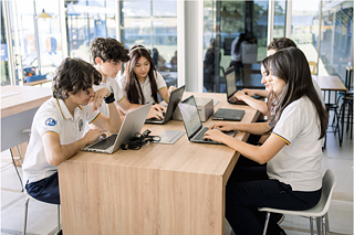 Mehrere Schülerinnen und Schüler sitzen um einen Holztisch und arbeiten an Laptops