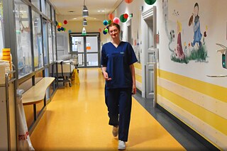 Ein junge Auszubildende läuft in blauer Arbeitskleidung den Gang eines Kinderkrankenhauses entlang.